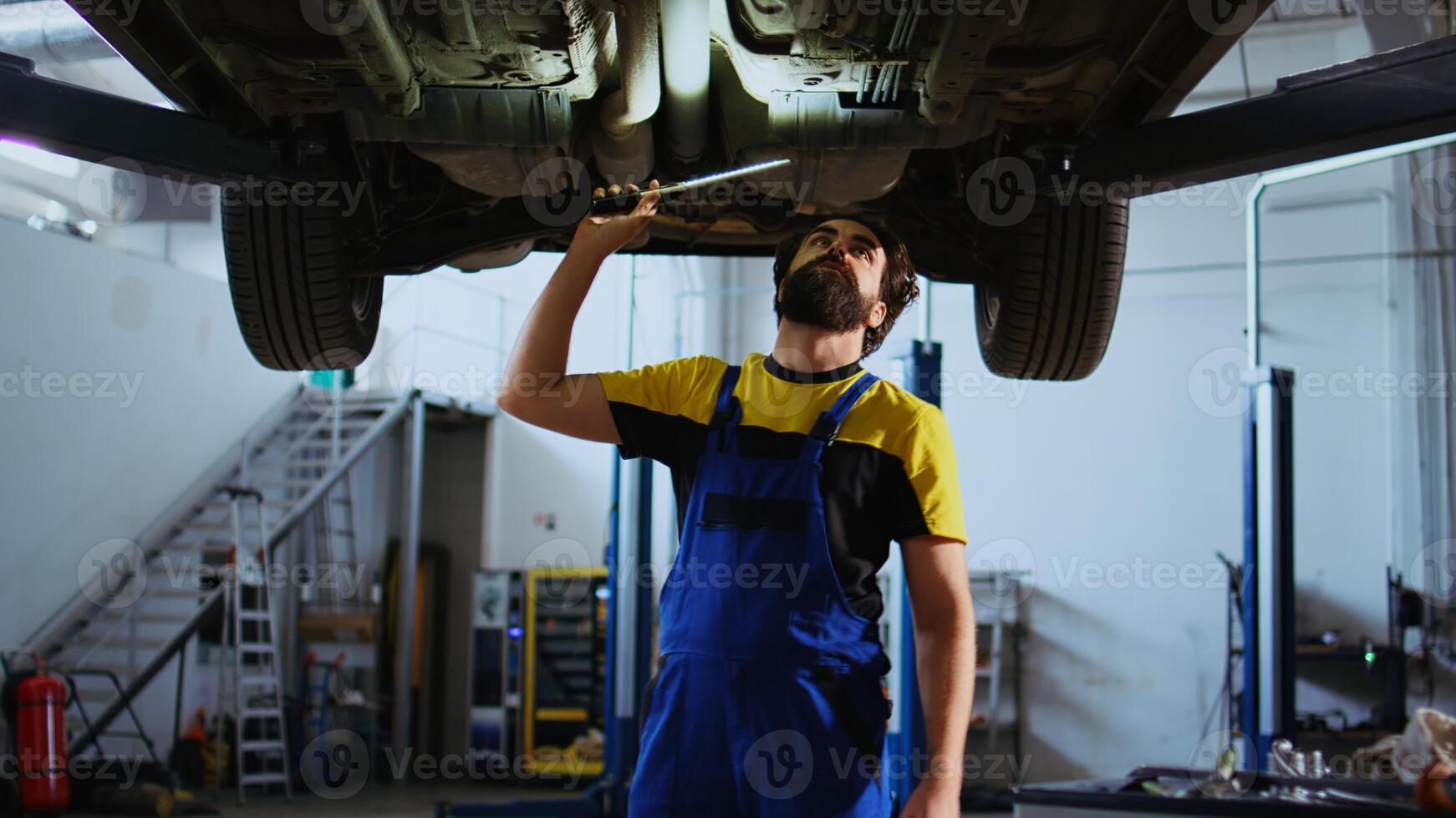 Meticulous serviceman working on suspended car in garage, checking components during routine maintenance. Man in auto repair shop walking underneath vehicle, surveying it using work light photo