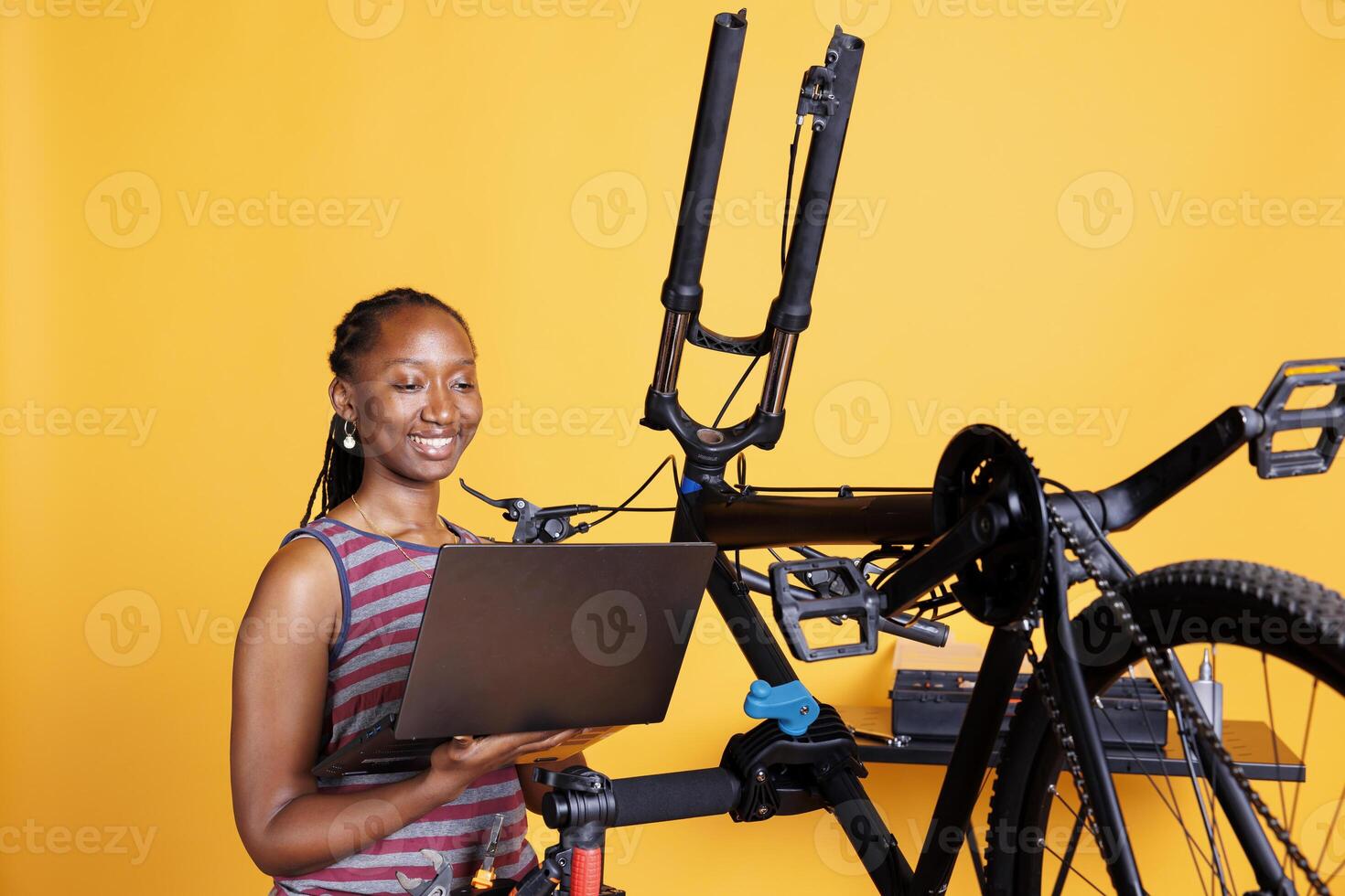 Female cyclist of african american ethnicity fixes damaged bike using toolkit and laptop to research solutions. Using minicomputer and professional equipment, black woman is repairing broken bicycle. photo