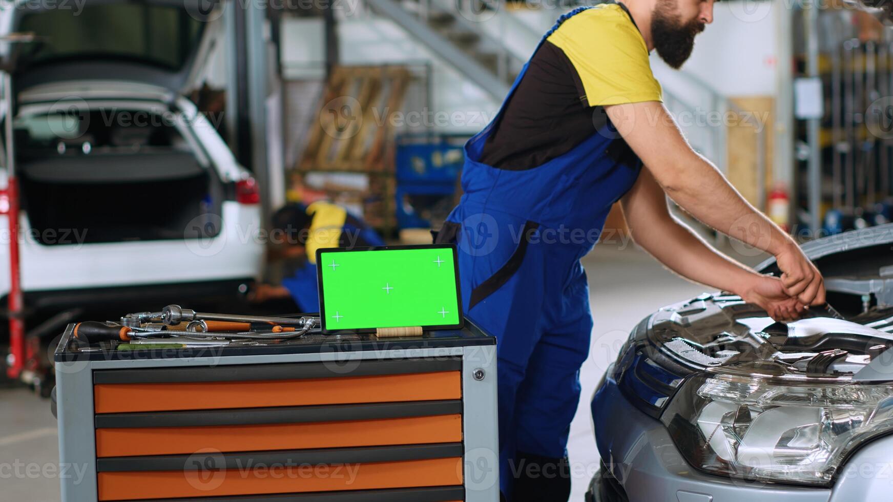 Panning shot of green screen laptop in garage workspace, sitting on work bench next to professional tools with mechanics in background working on cars, using torque wrench to fix them photo