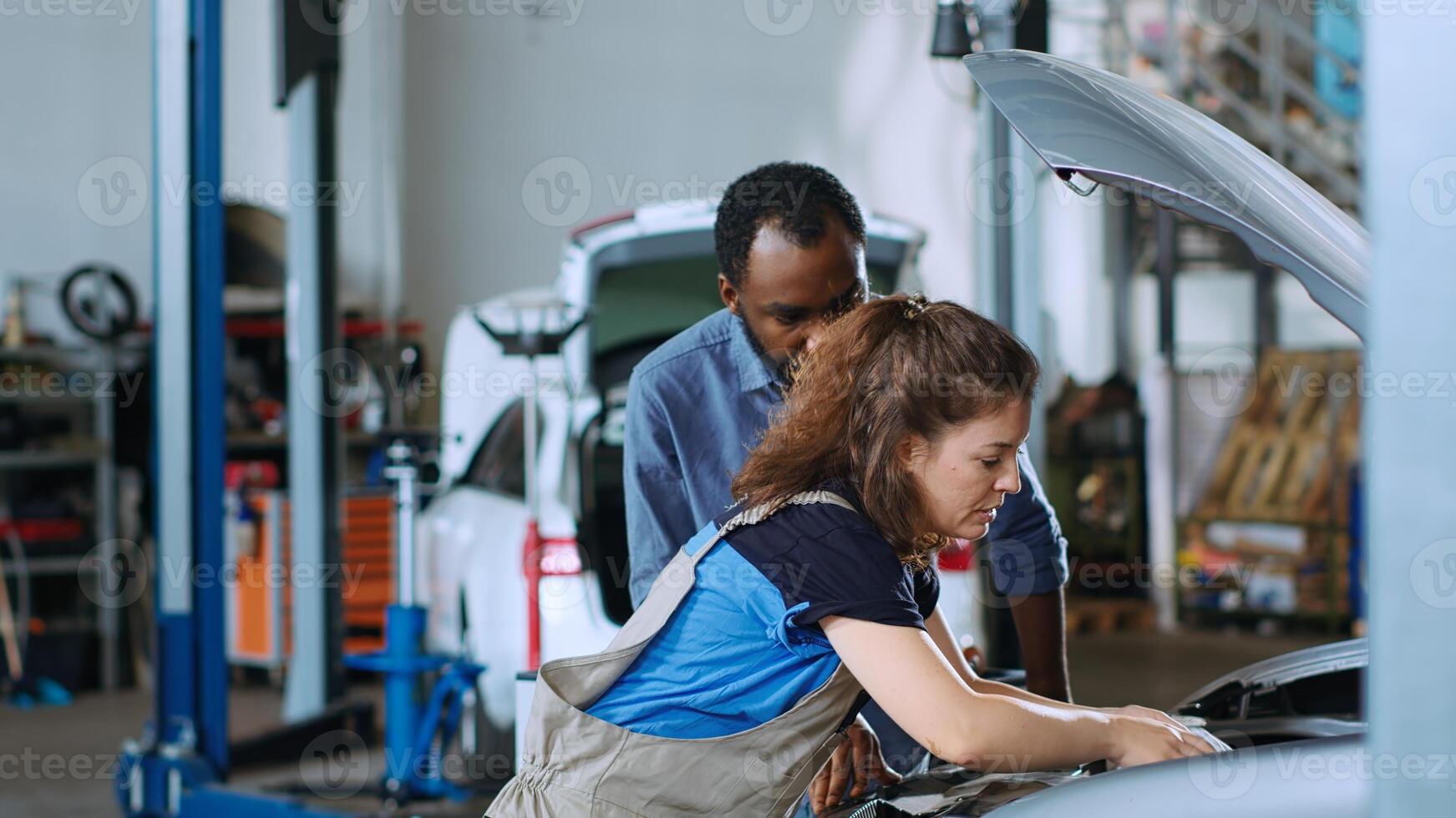 Mechanic in garage cleaning opened up vehicle after replacing oil while customer watches. Car service employee doing inspection on automobile, wiping leftover leakage and chatting with client photo