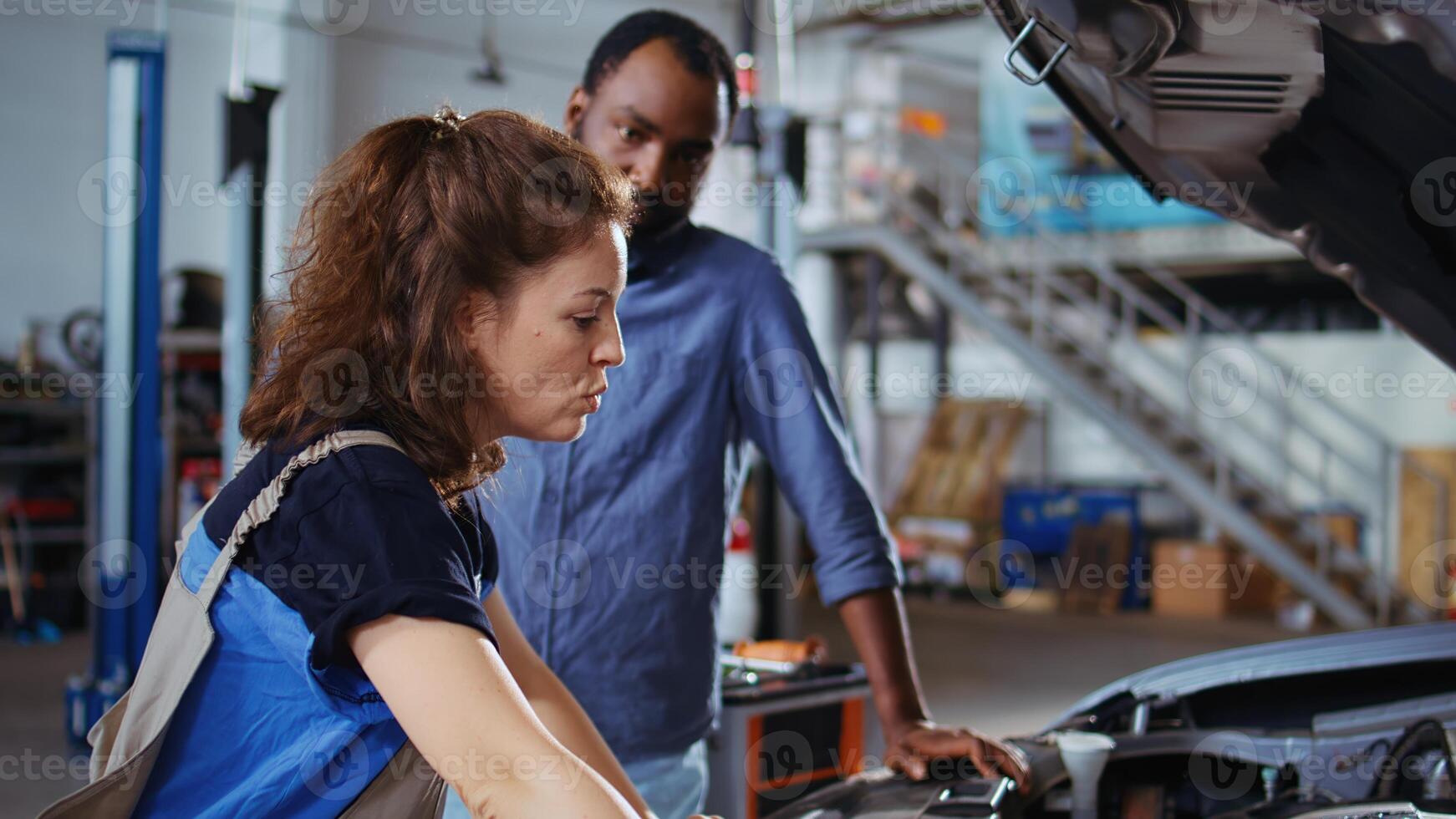 Trained engineer in garage finishing fixing car for customer, looking underneath vehicle hood to remove remaining oil leaks. Worker does routine parts cleaning on client automobile in auto repair shop photo