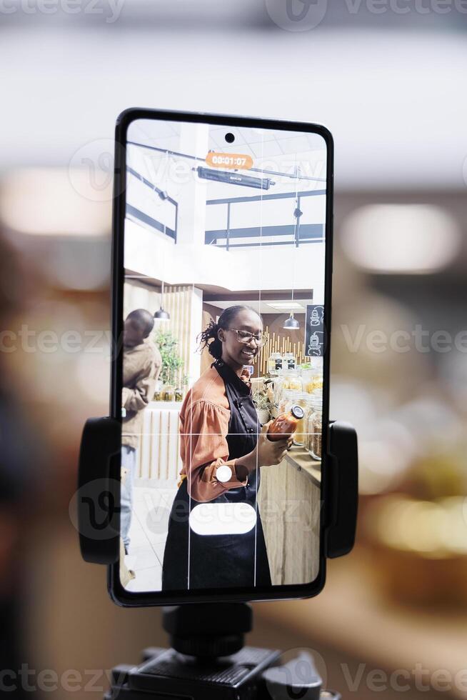African American store owner uses cell phone and selfie stick for vlogging, promoting sustainable, healthy and natural items. Organic, fresh food in glass jars, appealing to eco-conscious customers. photo