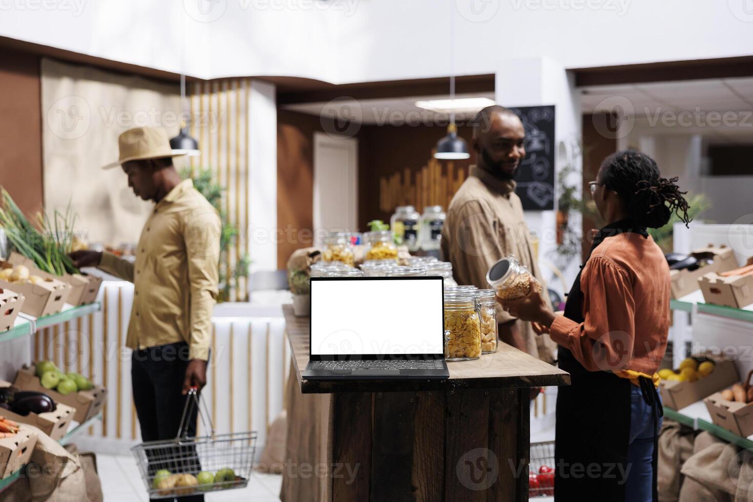 A laptop with an empty white screen display is set down on a wooden counter with customers looking around the store. In a modern food market, wireless computer has a blank chromakey mockup template. photo