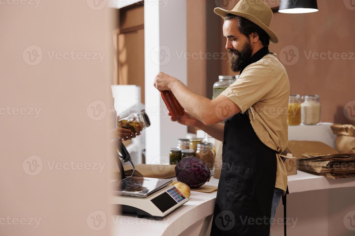 Caucasian male shopkeeper, who wears a stylish hat and black apron, stands behind the checkout counter holding a jar filled with pasta sauce. Vendor analyzing glass container at cashier desk. photo
