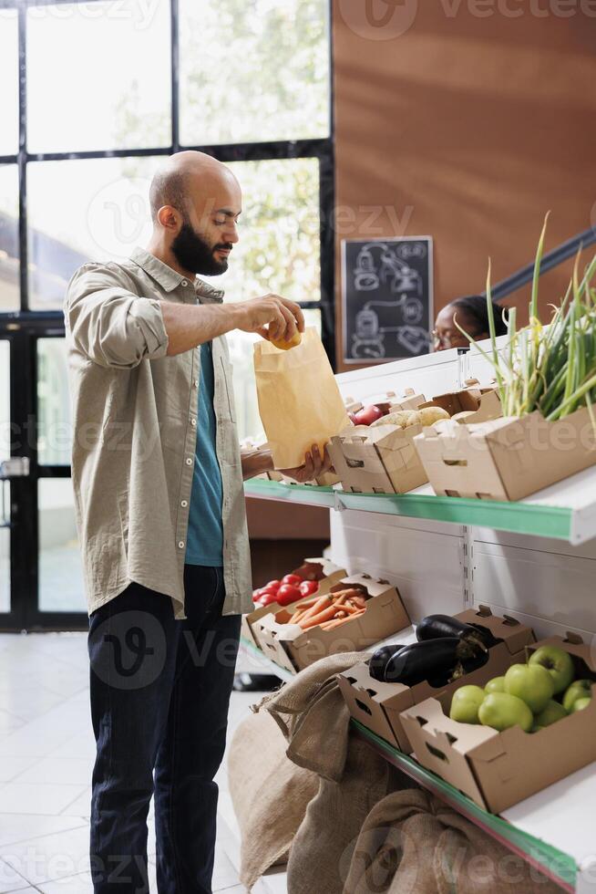 Middle eastern man explores modern eco friendly grocery store, browsing a variety of fresh, organic products with sustainable packaging. Male customer putting fresh potatoes in brown paper bag. photo