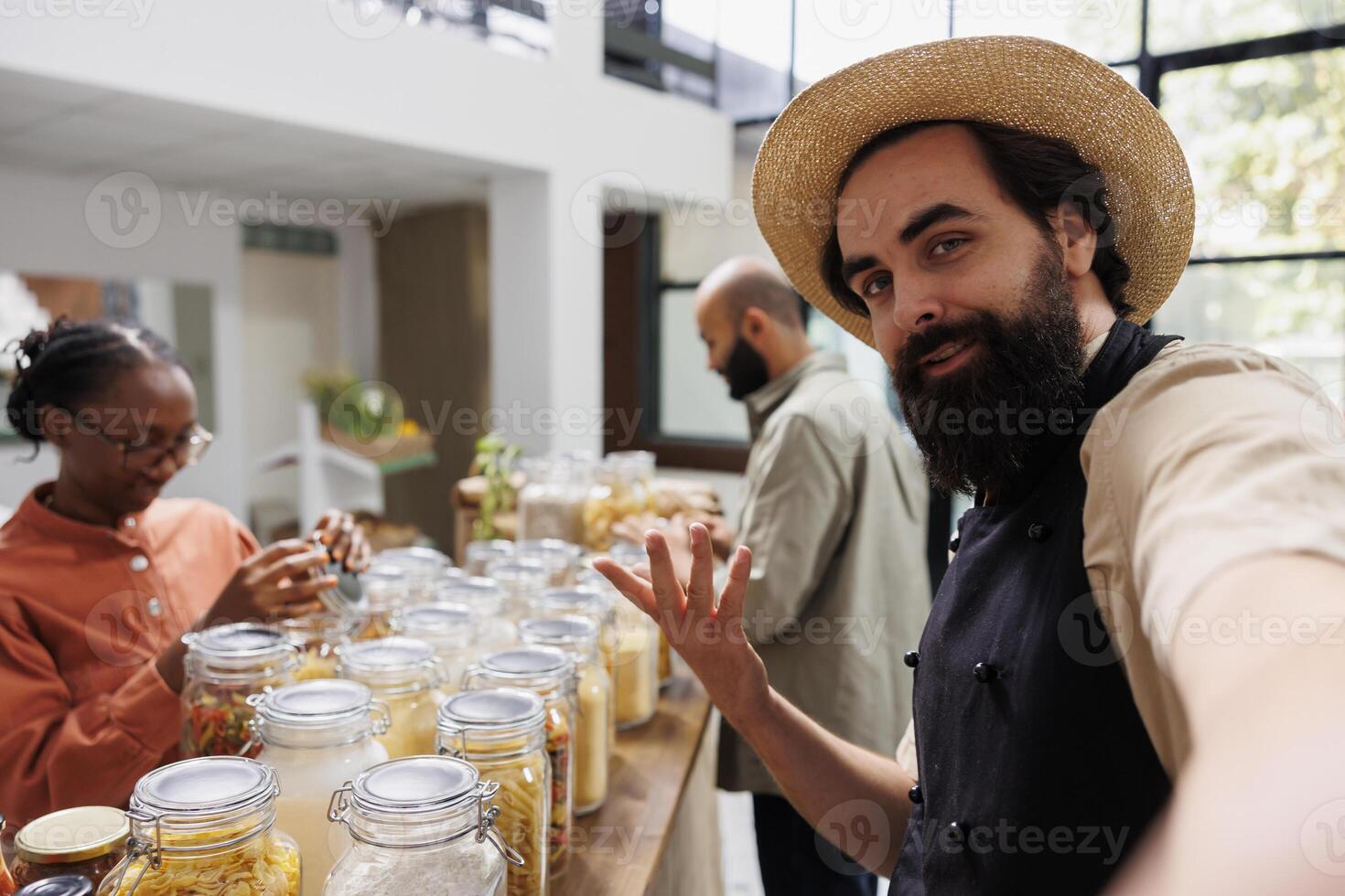 Bearded man wearing a hat and black apron gestures curiously to camera while black woman admires the contents in glass jar. Male client looks around in grocery store while vendor does vlogging. photo