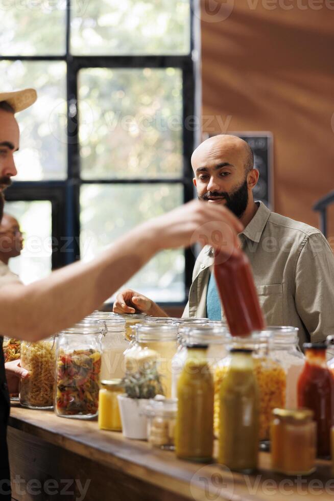 Middle Eastern customer looks at glass bottle filled with organic sauce being held by storekeeper. Local vendor providing male client with bulk natural sustainable products during an event. photo