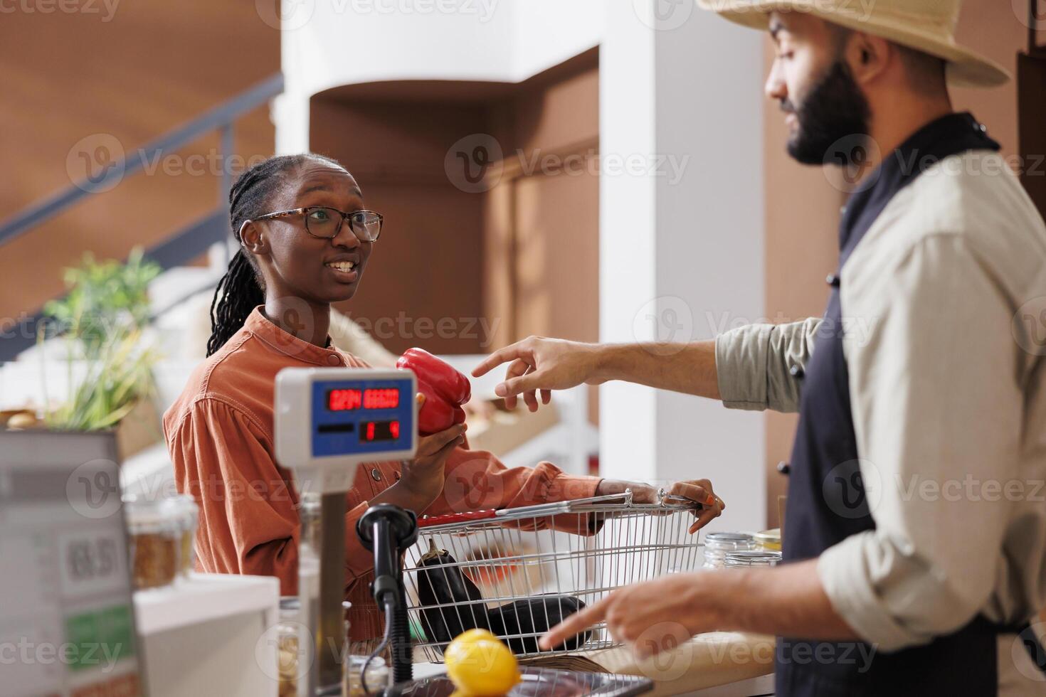 Black woman at checkout counter with a shopping basket filled with locally grown food products for vendor to weigh. Male cashier assisting african american client with her groceries. photo