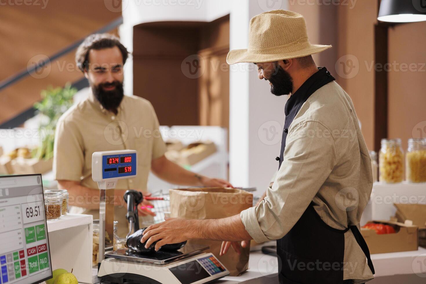 Middle eastern salesman being of service to caucasian customer at checkout counter. At cashier desk, male vendor wearing a hat while using digital scale to measure items for the young client. photo