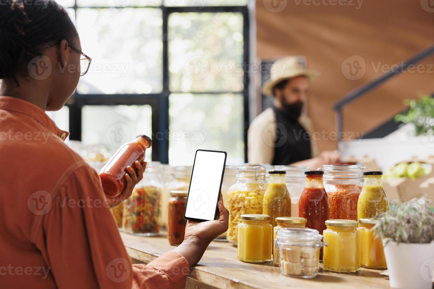 African american customer analyzes glass bottle filled with red sauce while also holding mobile device with white screen in grocery store. Black woman grasping cellphone with blank mockup template. photo