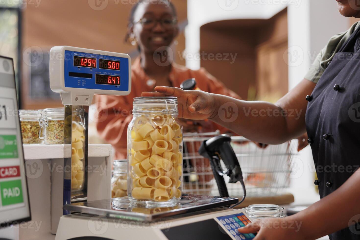 Black woman with spectacles waiting while vendor uses digital scale to measure glass container filled with fresh pasta. African american seller weighing jar of freshly made spaghetti for customer. photo