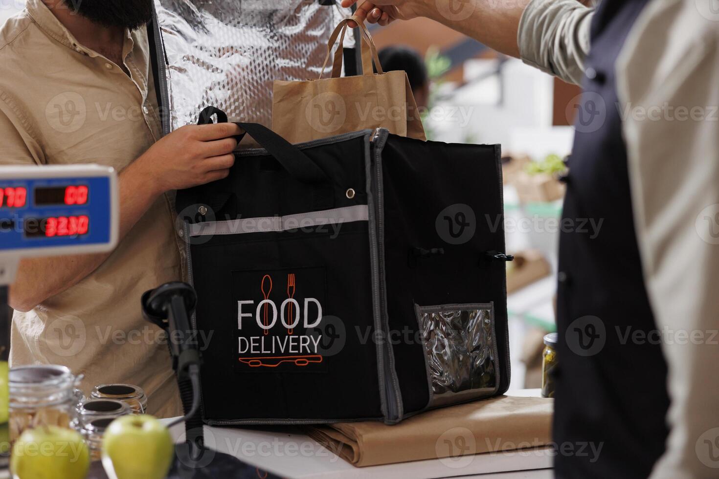 Close-up of the storekeeper placing takeaway into thermal bag for delivery man to send to customers with healthy living. Male courier collecting grocery order at counter of eco friendly store. photo