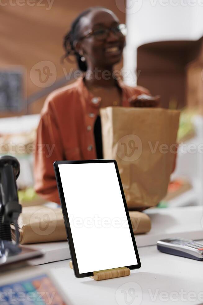 Closeup of digital tablet with blank white screen vertically placed on checkout counter at sustainable bio food market. Device displaying copy space mockup template while customer carries grocery bag. photo