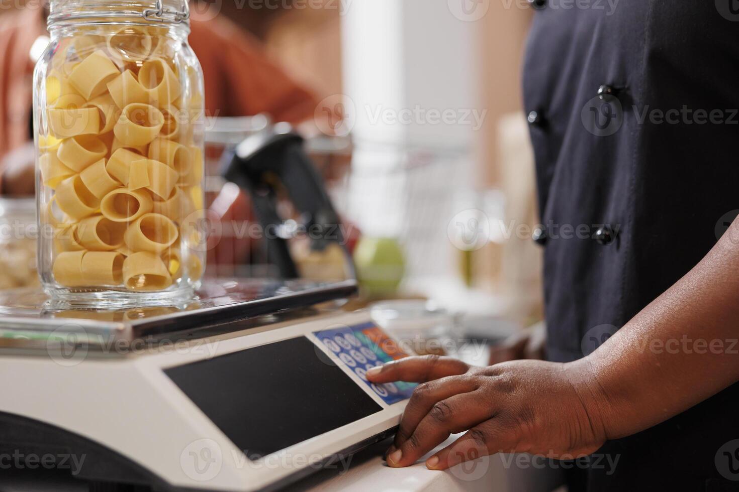 Up-close image capturing person using measuring scale, checking weight of container filled with organic bio goods. Black individual weighing glass jar of pasta at the checkout counter. photo