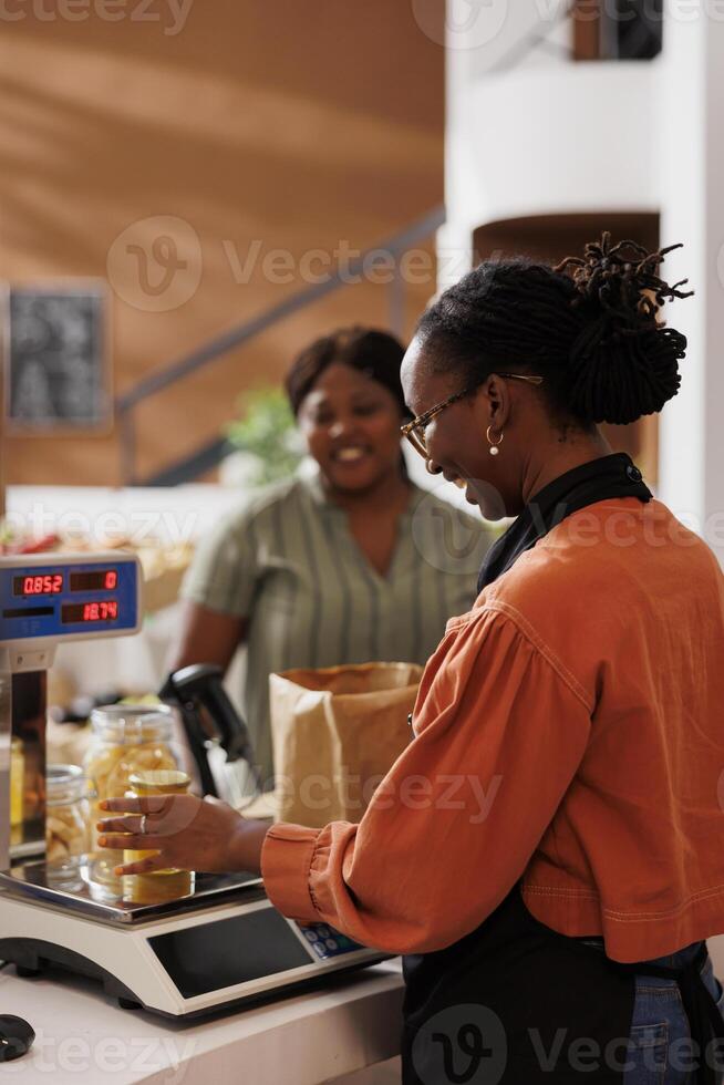 Female customer at a grocery store checkout counter with a smiling storekeeper weighing products on digital scale. Friendly vendor measuring weight of bio food items while client waits at desk. photo