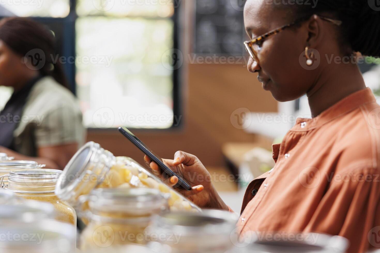 negro mujer con teléfono inteligente buscando para sano recetas para orgánico pasta mientras participación vaso envase. africano americano hembra cliente avaro tarro de espaguetis y surf el red en Teléfono móvil. foto