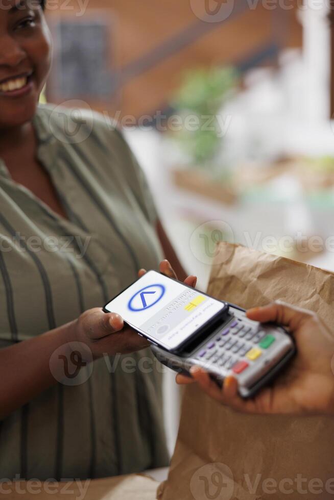 An African American woman smiles as she pays for freshly harvested organic produce using her mobile phone at a local marketplace. Convenient and eco-friendly shopping with contactless technology. photo