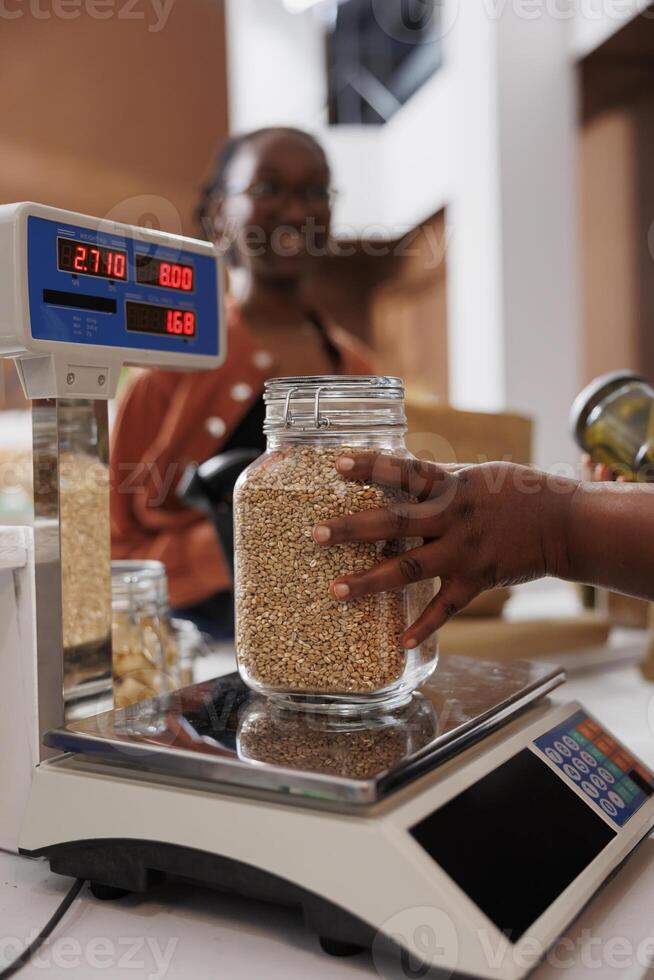 Closeup of black person assists customer with weighing fresh produce on digital scale at eco friendly store, promoting healthy, locally grown items. Seller measures sustainable additives free product. photo