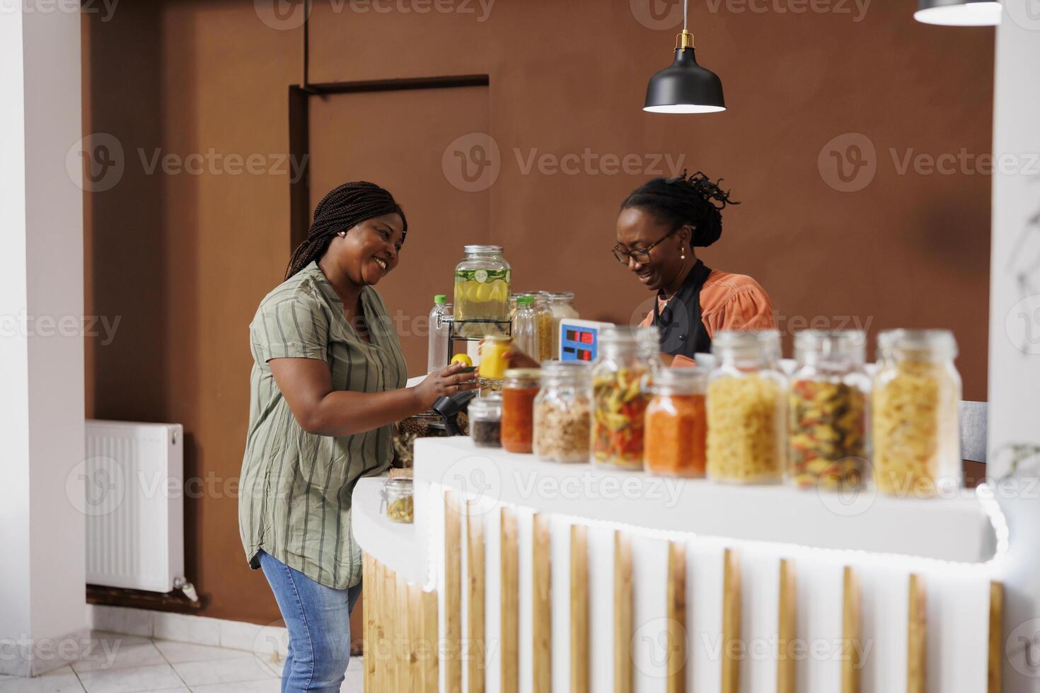 African American customer discusses eco friendly shopping with a store clerk at the checkout while handing over jars of cereal and honey for scanning. Black woman purchasing fresh bio food products. photo