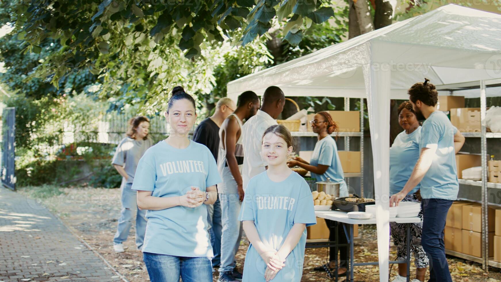 Portrait of a woman and daughter taking part in food bank program to fight hunger. Blue-shirted Caucasian volunteers are ready to help less fortunate while glancing into camera. Handheld shot. photo