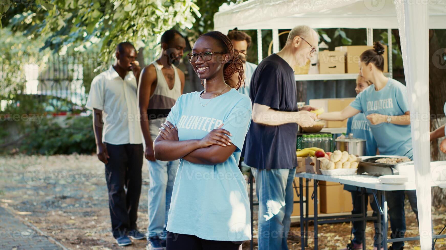 Side-view of black woman wearing glasses outside with arms crossed and eyes fixed on camera. Multiethnic volunteers supporting nonprofit initiative aimed at reducing hunger and aiding those in need. photo