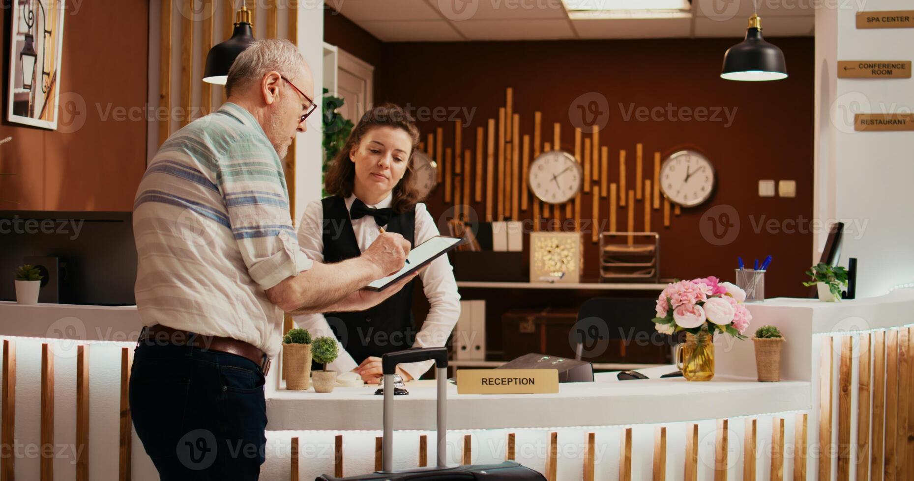 Old person signing check in forms at front desk, finishing with registration documents with employee at holiday resort. Tourist registering at all inclusive five star hotel, hospitality industry. photo