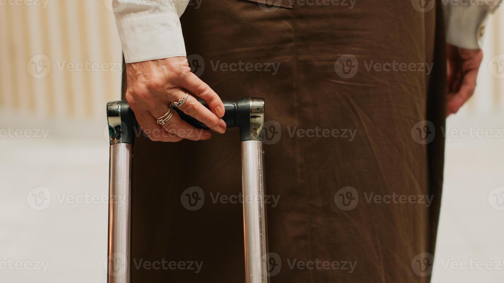 Older woman carries trolley bags at hotel reception, approaching front desk to do check in and start vacation trip. Traveller with luggage entering lobby, senior travel. Handheld shot. Close up. photo