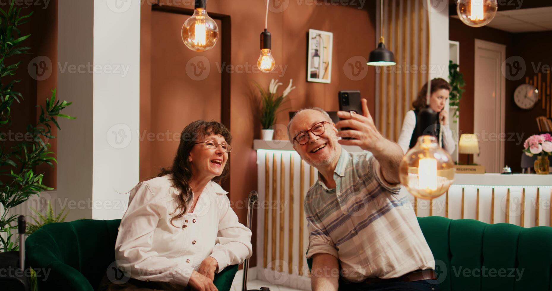 Senior couple talk on online videocall with nephews while travelling abroad, internet videoconference connection to present vacation plans. People passing time in hotel lounge area. photo