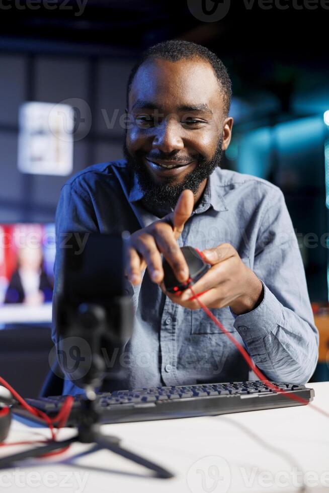 Smartphone on holder capturing footage of cheerful reviewer discussing with viewers about wired mouse. Influencer chats with fans during stream, filming computer peripheral review photo