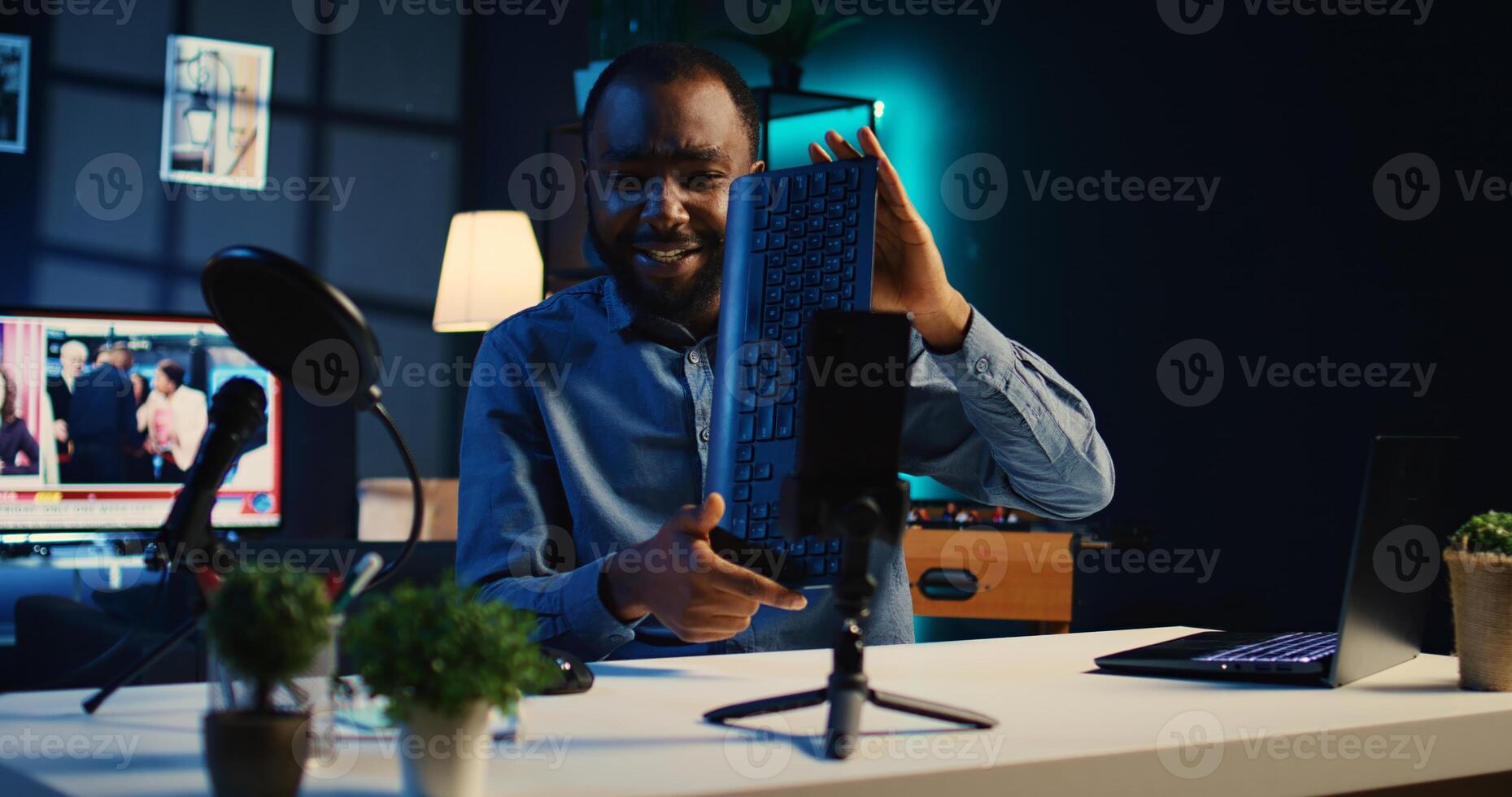 Content creator in dimly lit apartment studio records keyboard and mouse set review video for online streaming platforms. Cheerful man hosts internet show, presenting wireless computer peripherals photo