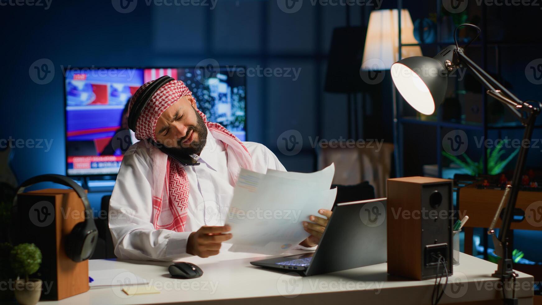 Muslim student at home in telephone call with school colleague discussing about finance class lesson and doing homework tasks together. Arab man at home desk looking over webinar notes photo