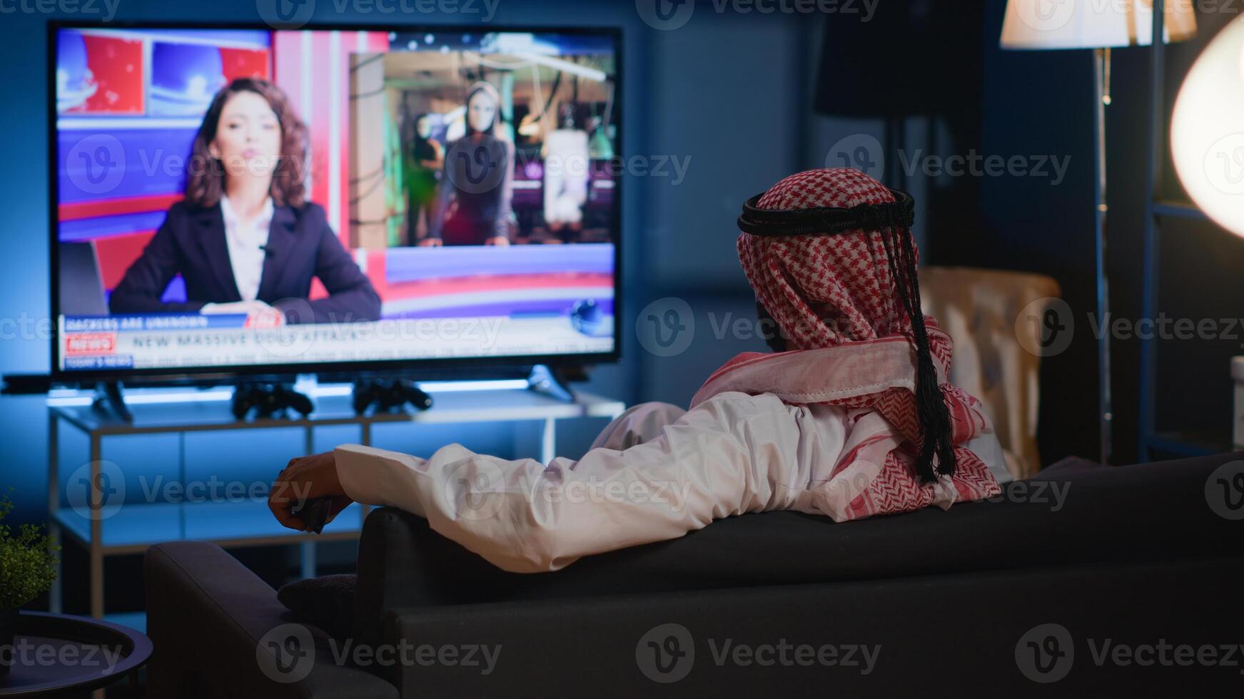 Arab man lounged on cozy sofa, watching news broadcast while relaxing at home after hard day at work. Muslim person chilling on couch, looking at TV channel showing current events photo