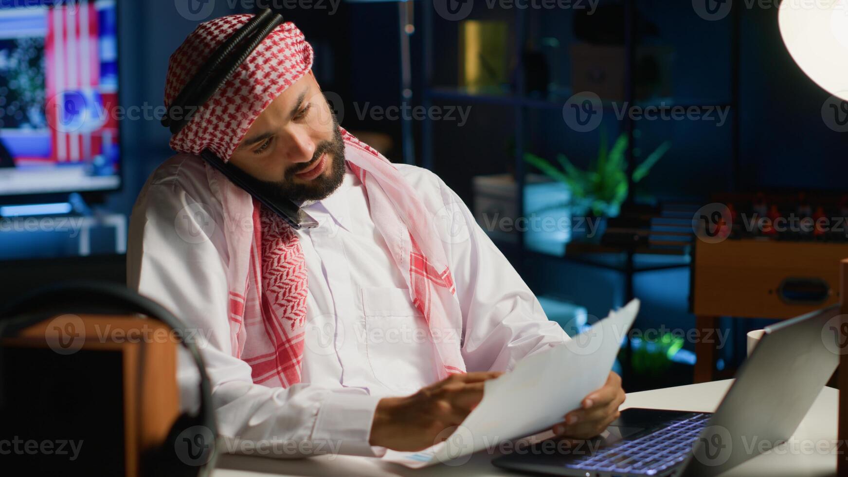 Arabic employee having discussion with coworker in phone call while remotely working from cozy home office. Teleworker talking with colleague using smartphone, discussing paperwork photo
