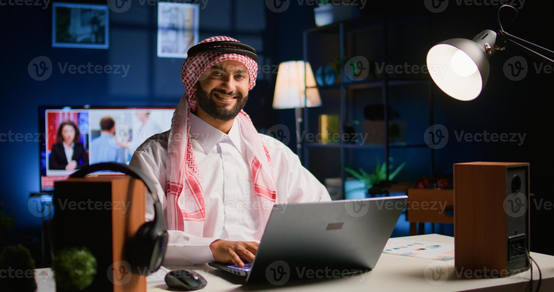 Happy arabic teleworker answering job emails in stylish apartment. Cheerful muslim employee remotely working, typing data on laptop with opened tv as background noise, handheld camera shot photo