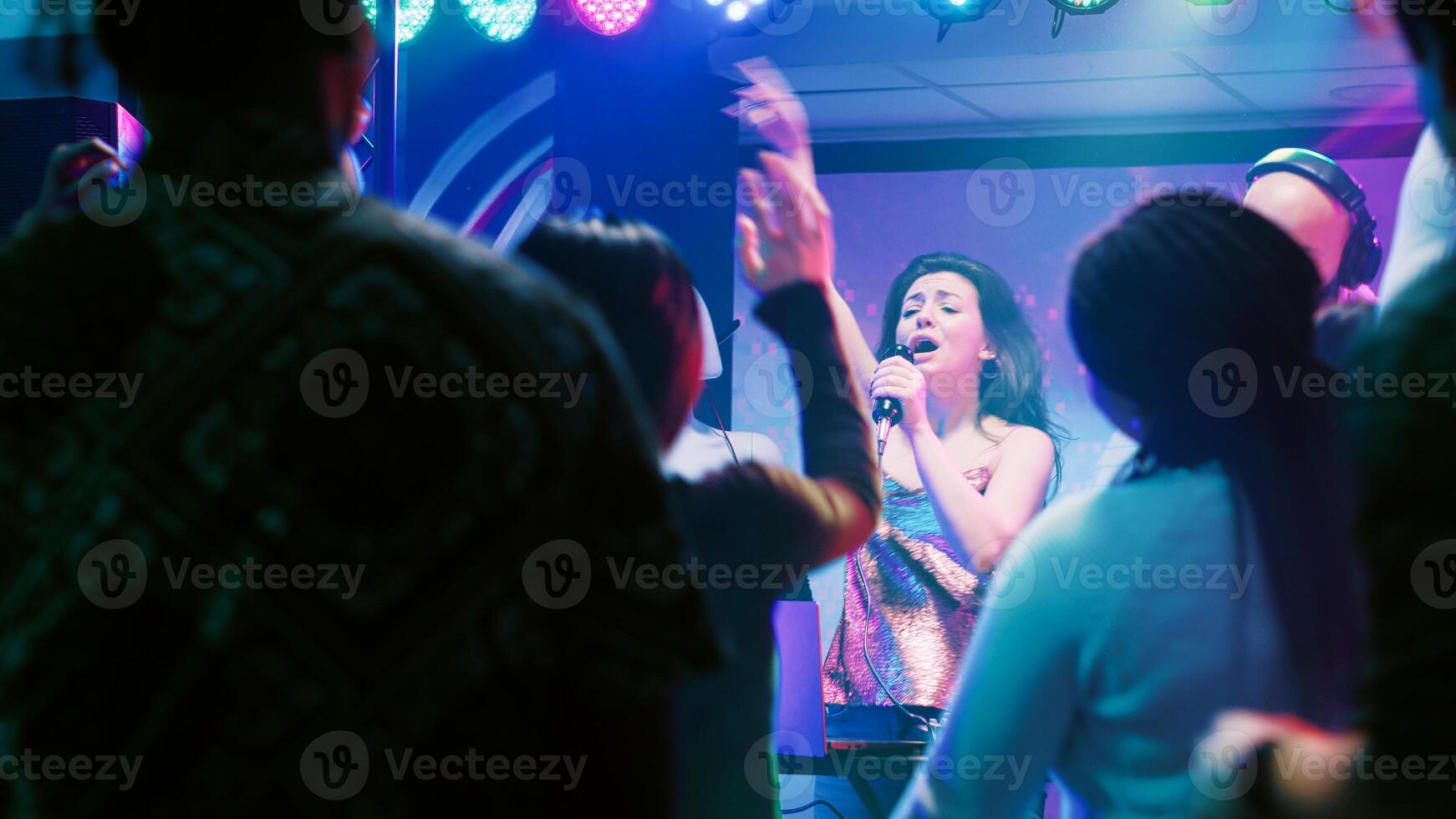 Confident woman doing karaoke on stage, singing songs with male DJ in front of people at nightclub. Happy girl having fun with live perfromance next to audio mixing station in club. photo