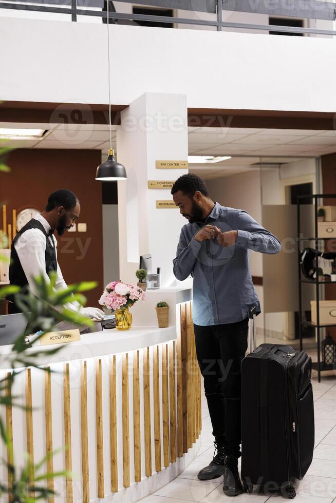 Young African American man tourist arriving to hotel, standing alone with suitcase at reception counter. Black millennial guy traveler checking in at front desk, waiting for room key photo