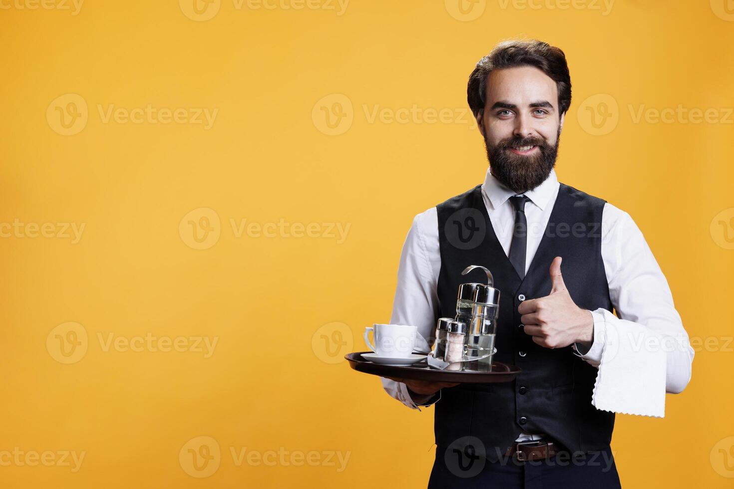 Confident waiter shows thumbs up on camera, showcasing positive approval symbol while he carries coffee on platter. Young man butler does okay sign, being in agreement at restaurant job. photo