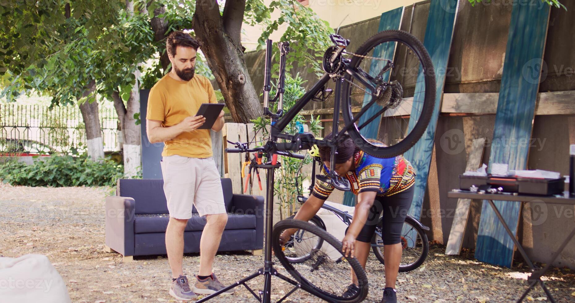 caucásico hombre en pie con inteligente dispositivo mientras africano americano mujer desmantela dañado bicicleta rueda. Pareja utilizando digital tableta para investigando de bicicleta ajustes para verano mantenimiento. foto