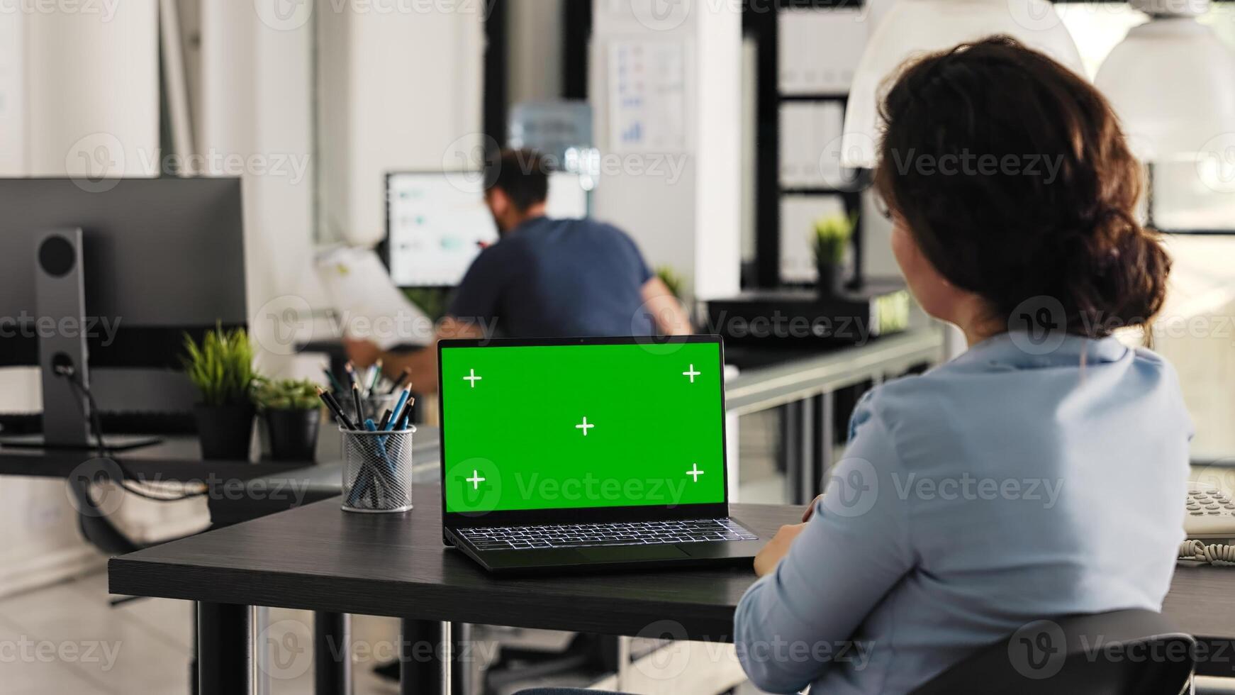 Businesswoman works on laptop with greenscreen, looking at blank chromakey display in coworking space. Young adult examining pc screen with isolated mockup template, e commerce tasks. Handheld shot. photo