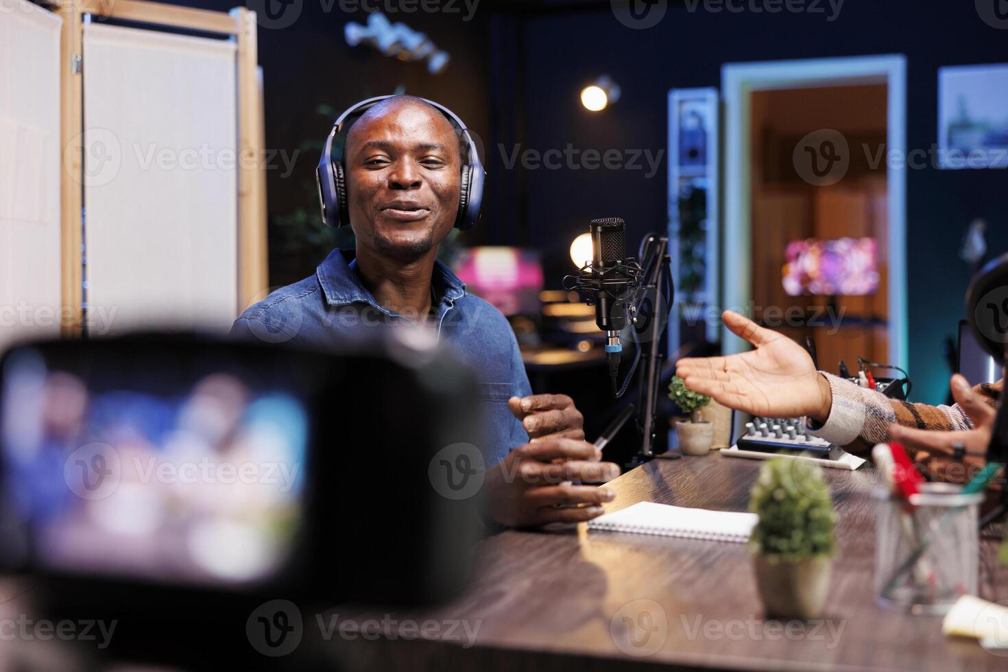 Cheerful influencer looks at camera mounted on tripod, announcing a guest on his show. African American guy wearing headphones using professional recording equipment to film video material. photo