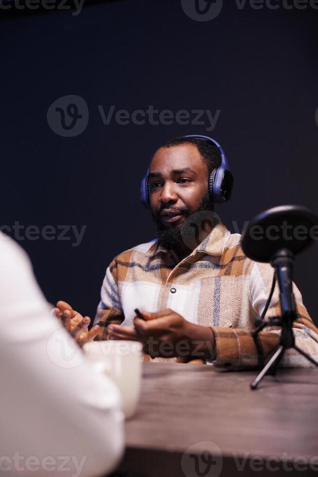 Black male host interviewing a person at home, using modern microphone and wireless headphones for his online talk show. African american man talking with an individual recording a podcast episode. photo