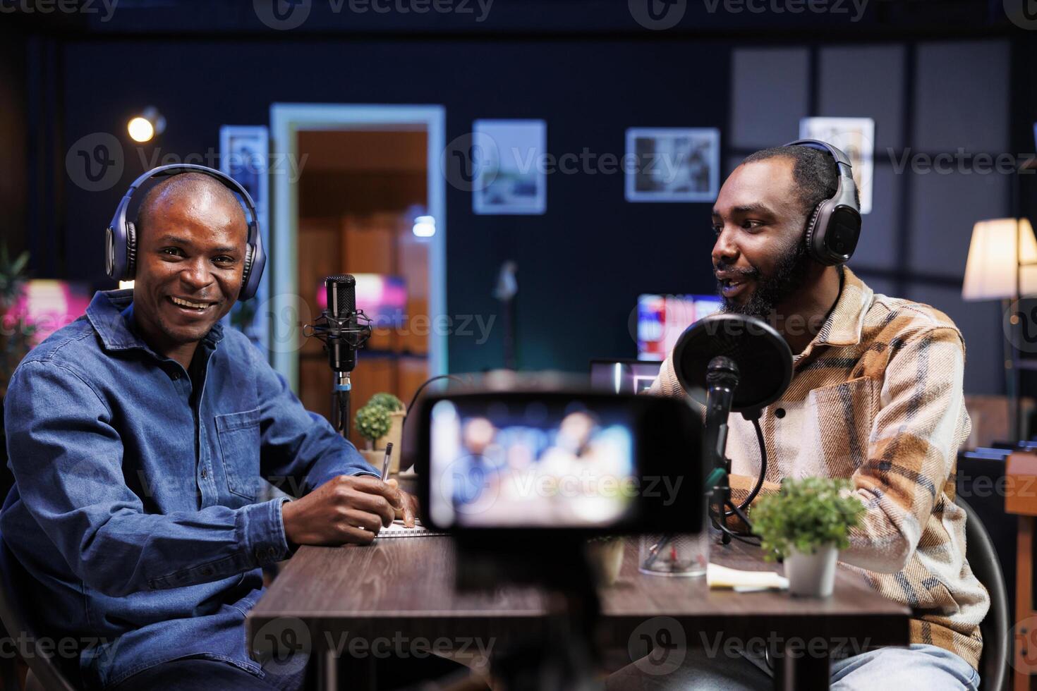 Smiling journalist introduces a guest on his show while facing the camera and speaking into microphone. Black men with headphones are recording videos using a camera mounted on a tripod. photo