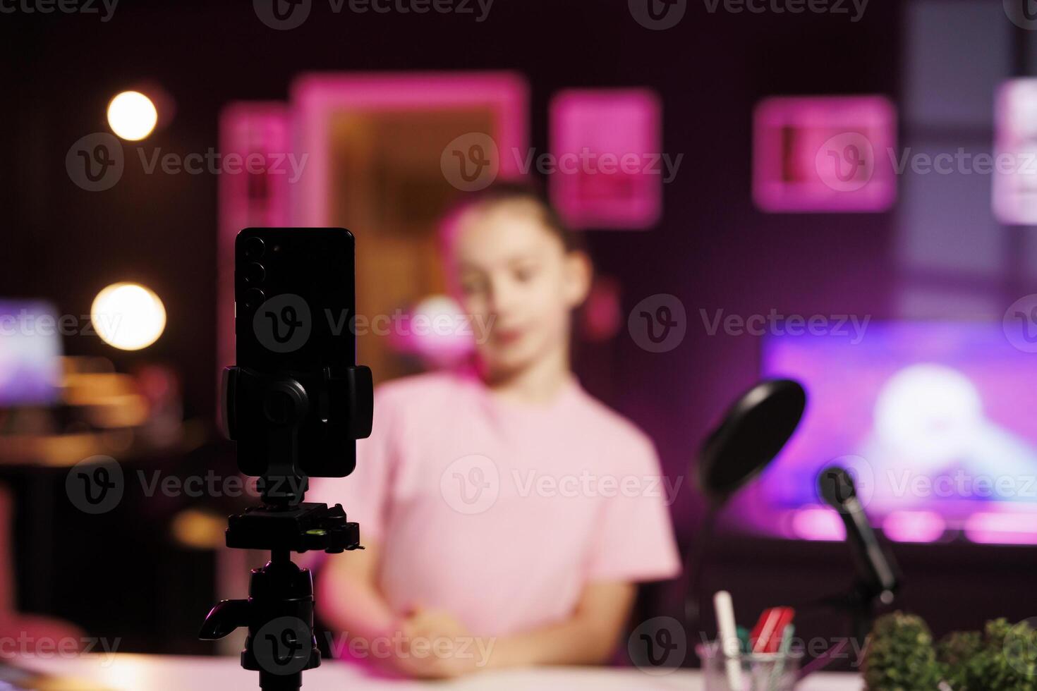 Focus on smartphone on tripod used by little girl shooting product unboxing for streaming channel on online platforms. Child in blurry background warmly welcoming fans, filming reviews photo