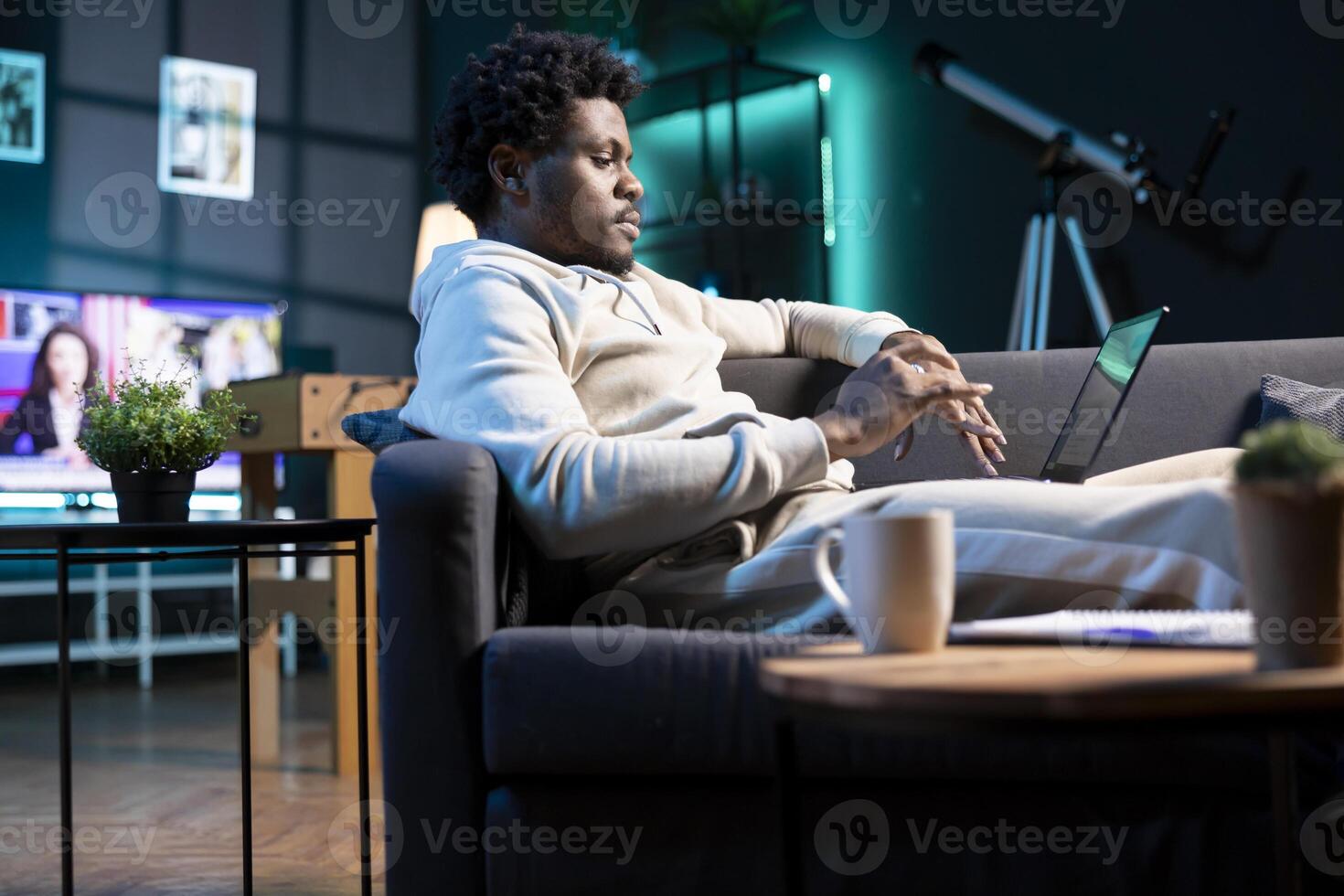 Self employed man inputting data on laptop while working from home, looking at screen. BIPOC person lying on couch, remotely performing tasks in nice stylish warm apartment living room photo