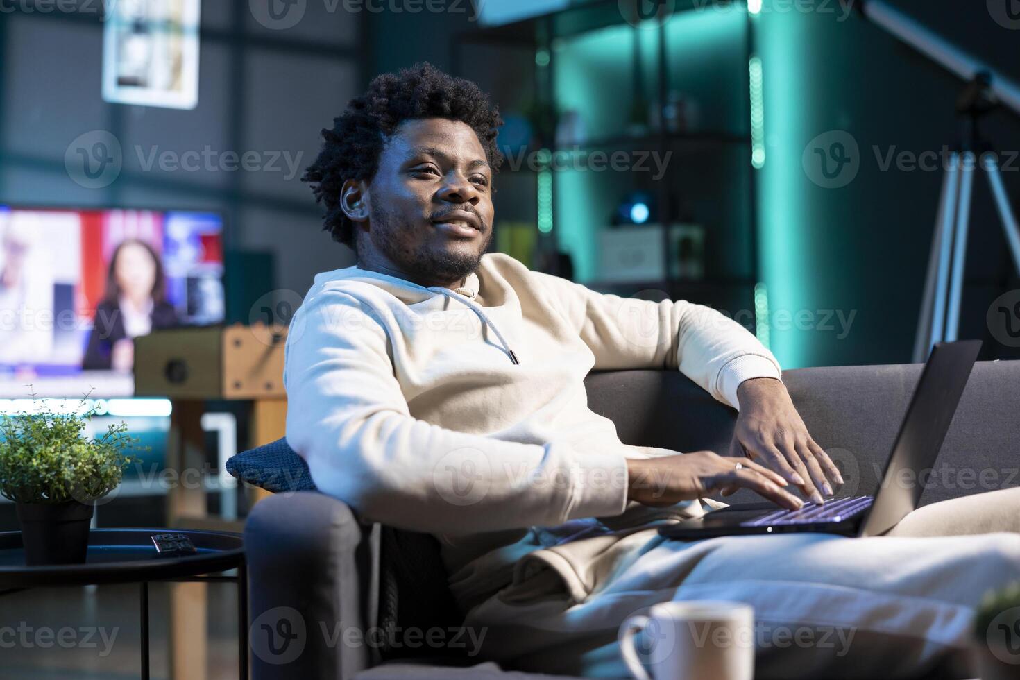 Smiling teleworker answering job emails in stylish apartment, happy about finishing work soon. Cheerful african american employee remotely working, typing data on laptop with tv background noise photo