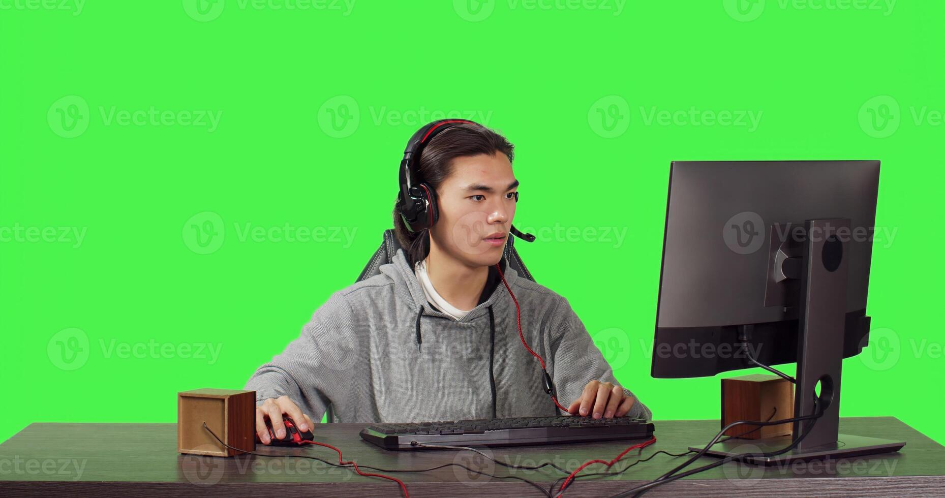Young gamer sitting at computer desk and playing web based video games with people, using modern gadgets to have a good time. Asian person competing in rpg contest over greenscreen. photo