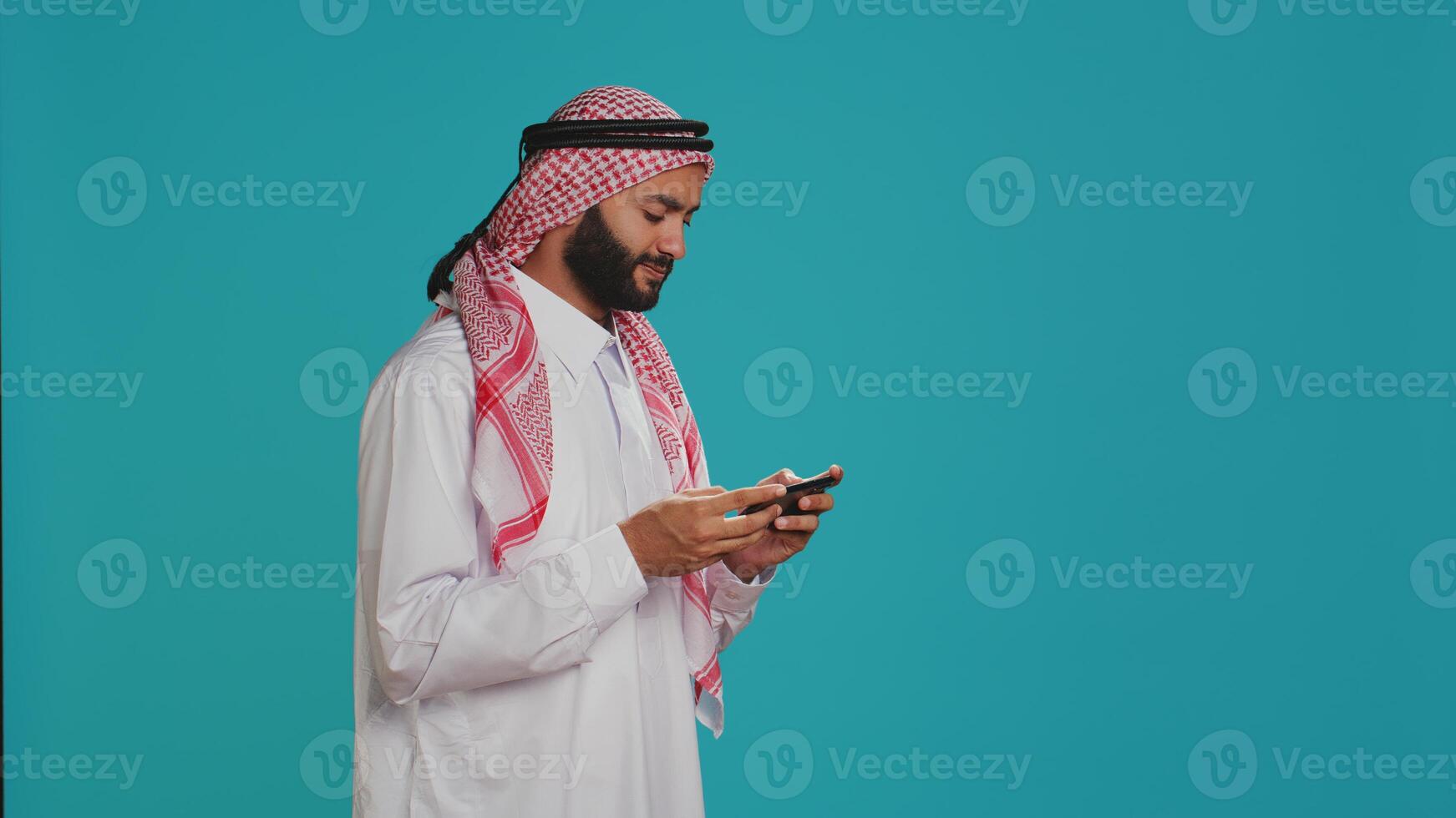 Muslim man wears traditional attire and playing videogames on smartphone, standing over blue background. Arab person enjoying mobile gaming competition on phone app, entertainment. photo