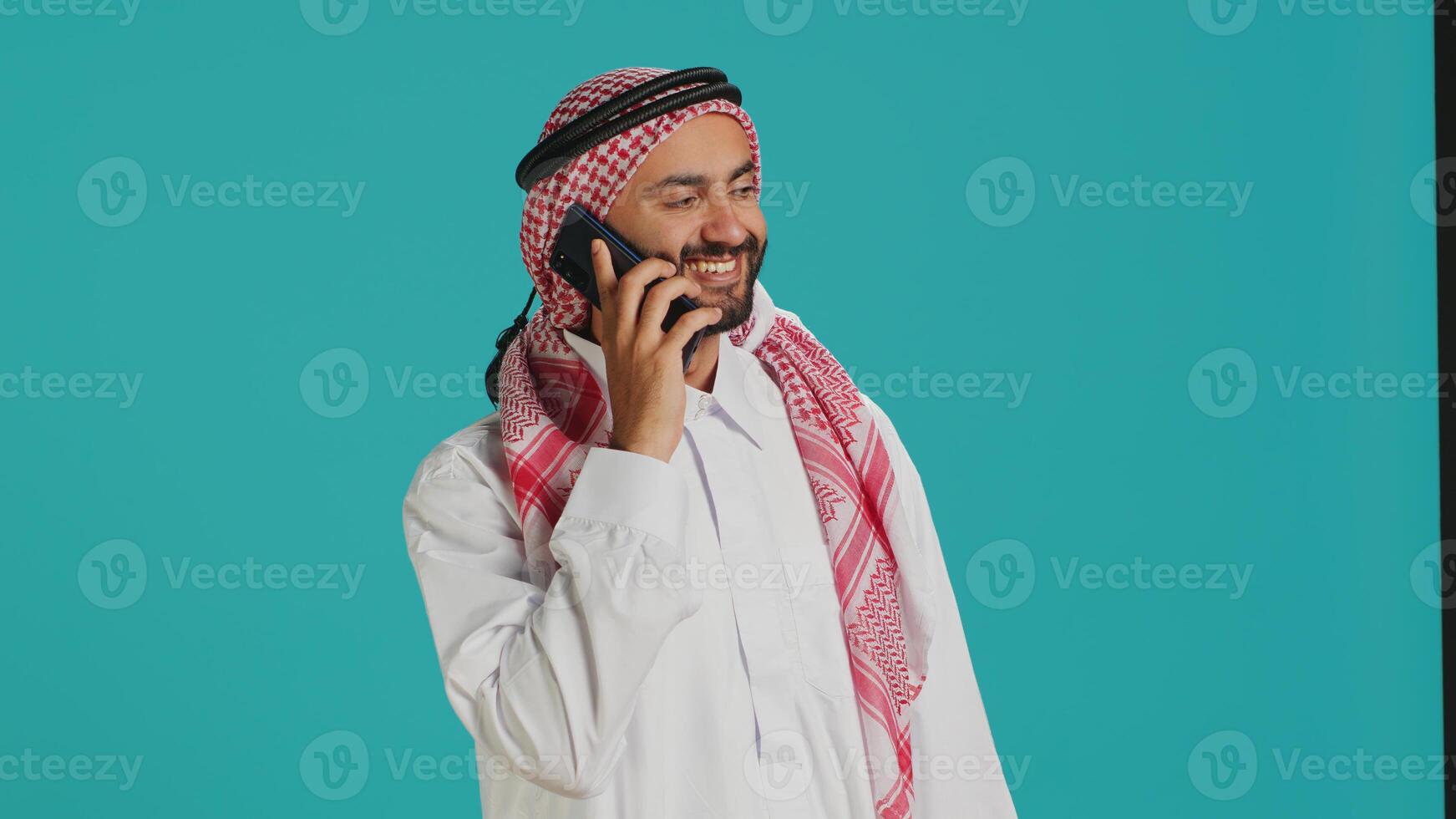 Muslim man in traditional attire texting on mobile phone while posing proudly in studio. Middle eastern adult dressed in thobe and checkered headscarf, sending online messages on phone. photo