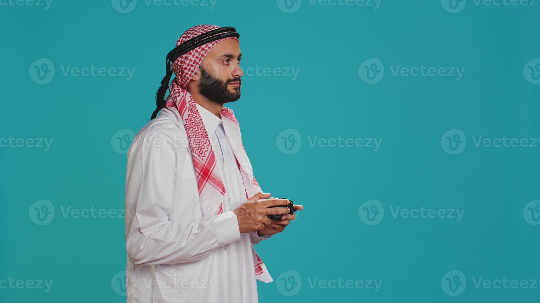 Modern man playing games with controller, having fun with video gaming competition over blue studio background. Islamic player in traditional arab clothing competes in tournament. photo