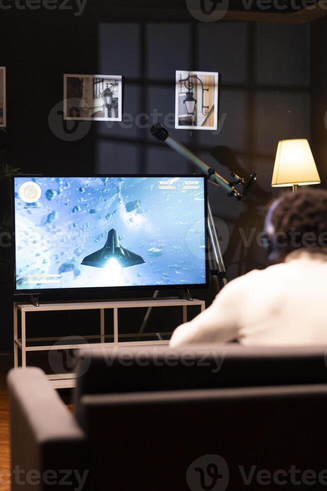 Gamer in front of widescreen smart TV watching SF videogame gameplay. African american man wearing headphones in home theatre to enjoy gaming content online displayed on television set photo